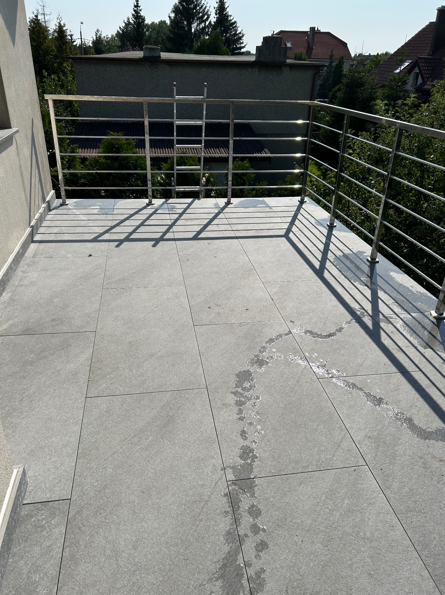 Wykończony balkon z szarą, dużą płytką i metalową balustradą. Widoczne ślady po myciu płytek. W tle zieleń drzew i zabudowa miejska.