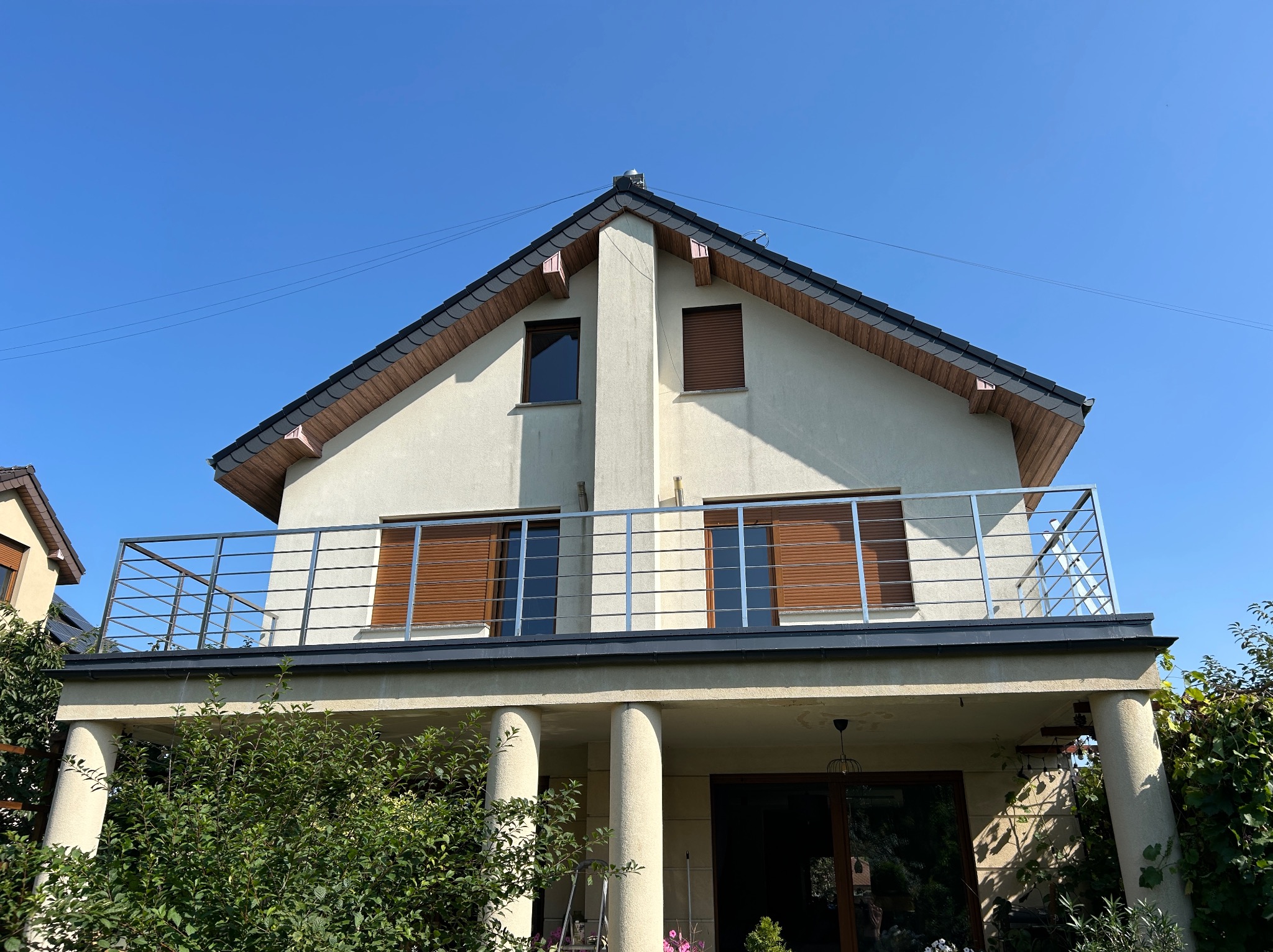 Dwupiętrowy dom z balkonem i brązowymi okiennicami, widziany z dołu na tle błękitnego nieba. Stalowa balustrada balkonu i kolumny podtrzymujące balkon.
