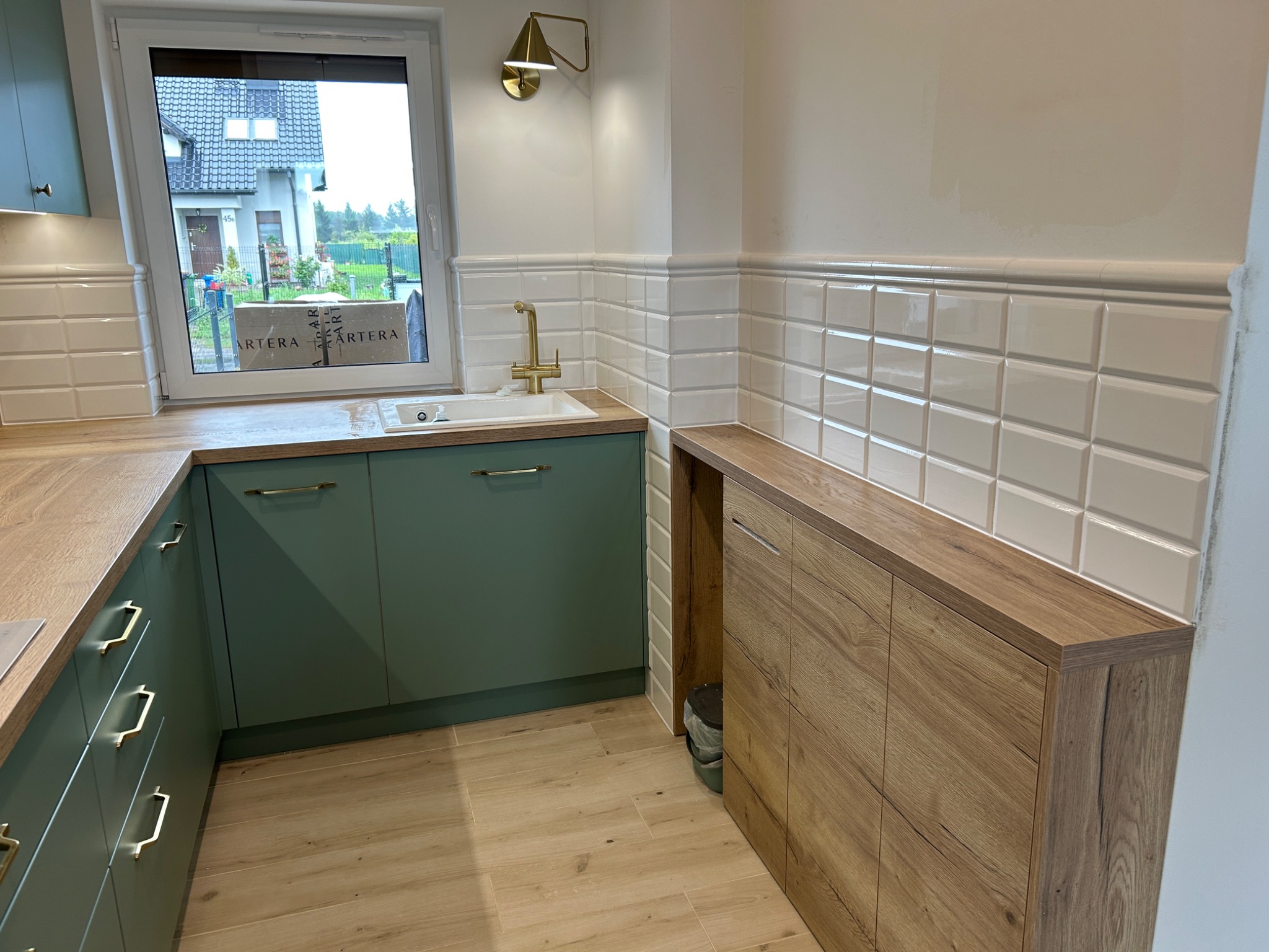 Wykończona kuchnia z zielonymi szafkami i złotą armaturą. Ściany zdobią białe, wypukłe płytki w stylu retro. Jasne drewno na podłodze i blacie dodaje ciepła.