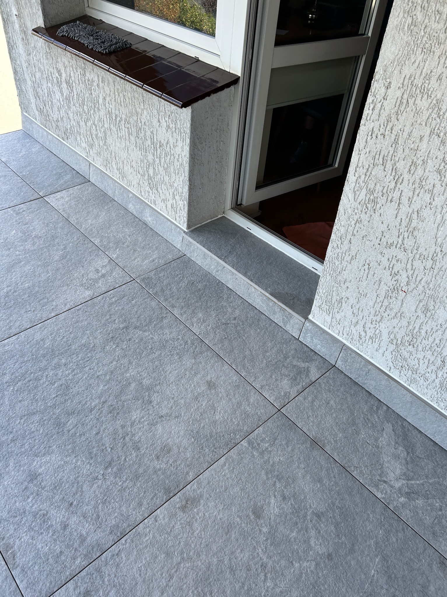 Wykończony balkon z szarymi płytkami podłogowymi i oknem z brązowym parapetem. Ściany z jasnym tynkiem strukturalnym. Widok z góry, ukośny kadr.