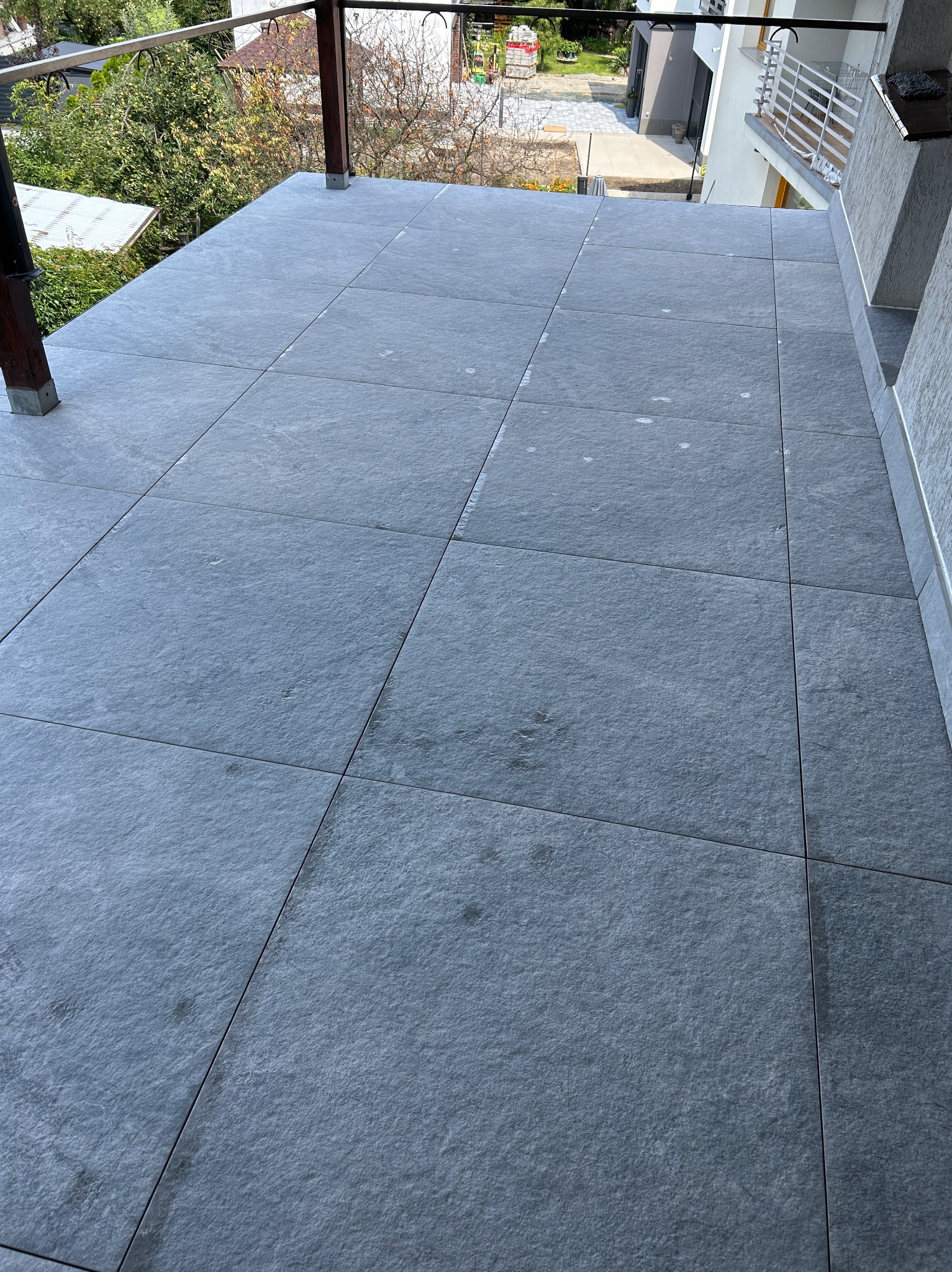 Wykończony balkon z szarymi, kwadratowymi płytkami. Widok z góry, ukazujący równo ułożoną podłogę i elementy konstrukcyjne balkonu. Minimalistyczny design.