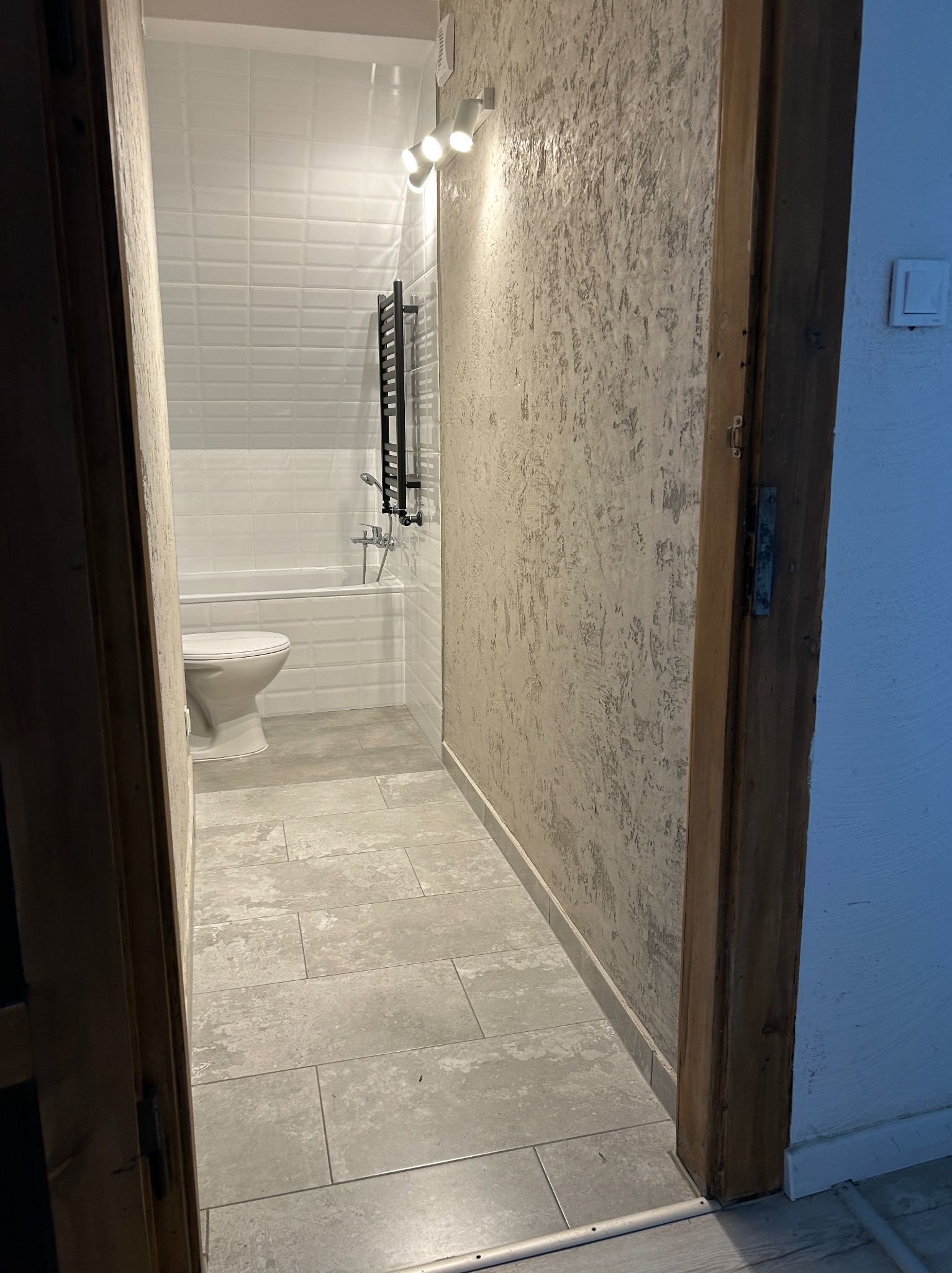 Wykończona łazienka z wanną, toaletą i grzejnikiem drabinkowym, widoczna przez otwarte drewniane drzwi. Ściany wykończone w bieli i szarości, płytki podłogowe w odcieniach szarości.