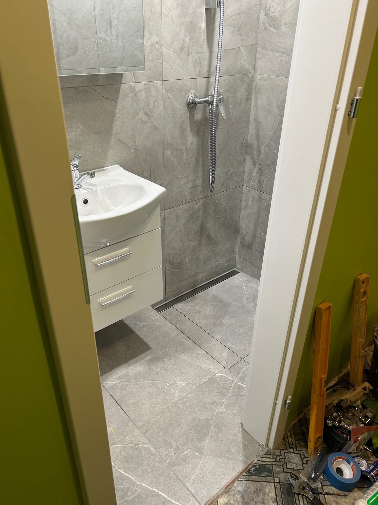 Wykończona łazienka z szarymi płytkami, kabiną prysznicową i białą szafką z umywalką, widok zza futryny drzwi. Minimalistyczny design, nowoczesne wykończenie.