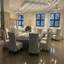 Elegancko nakryte okrągłe stoły z białymi obrusami i pokrowcami na krzesłach w jasnej sali bankietowej, dekoracja kwiatowa na stole, widok na okna i oświetlenie sufitowe.