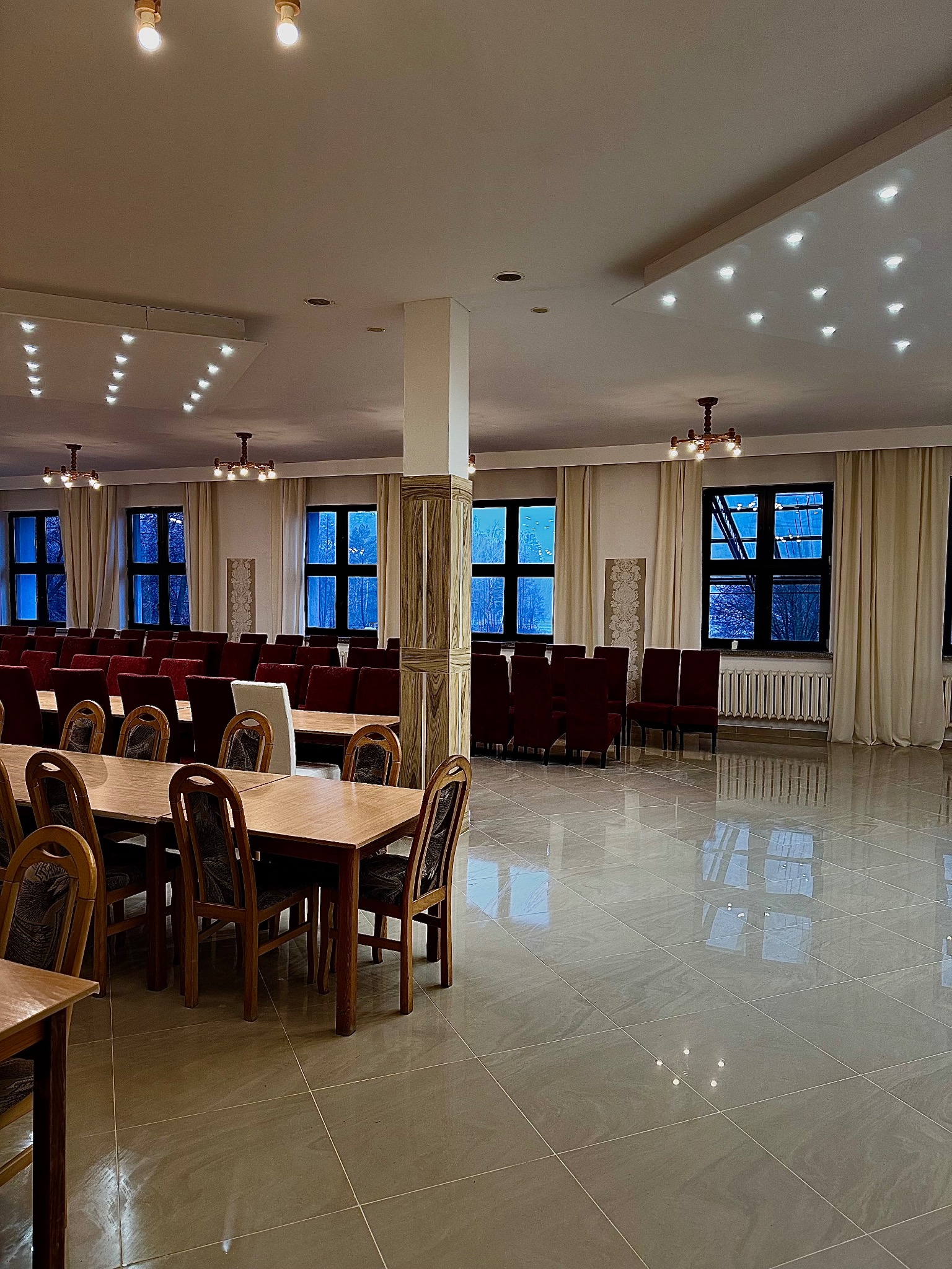 Sala bankietowa z drewnianymi stołami i krzesłami z bordową tapicerką, oświetlona sufitowymi lampami i żyrandolami, z widokiem na zimowy krajobraz za oknami.