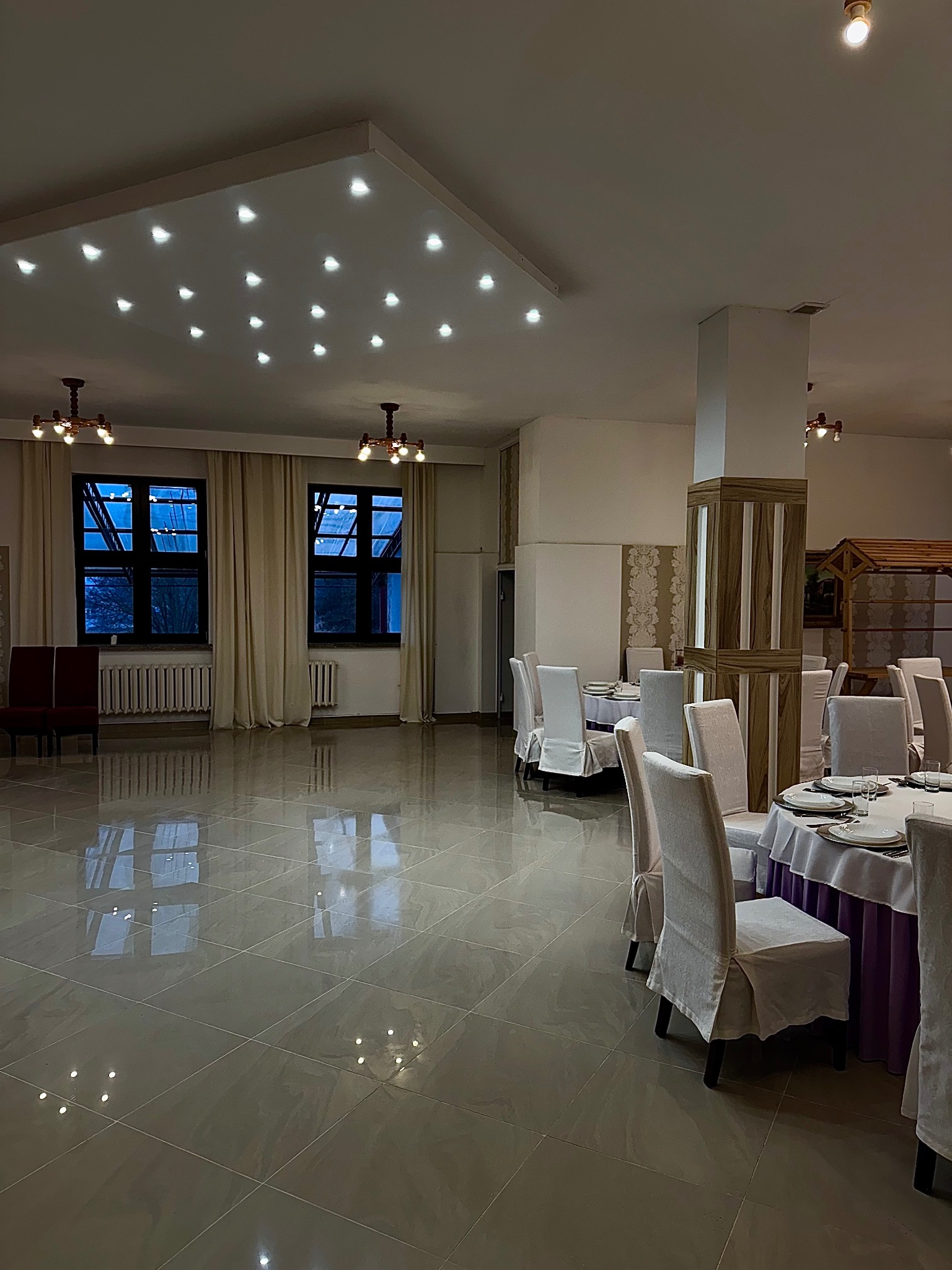 Sala bankietowa z okrągłymi stołami nakrytymi białymi obrusami z fioletowymi akcentami, krzesła z białymi pokrowcami, parkiet odbijający światło, sufit z panelami oświetleniowymi.