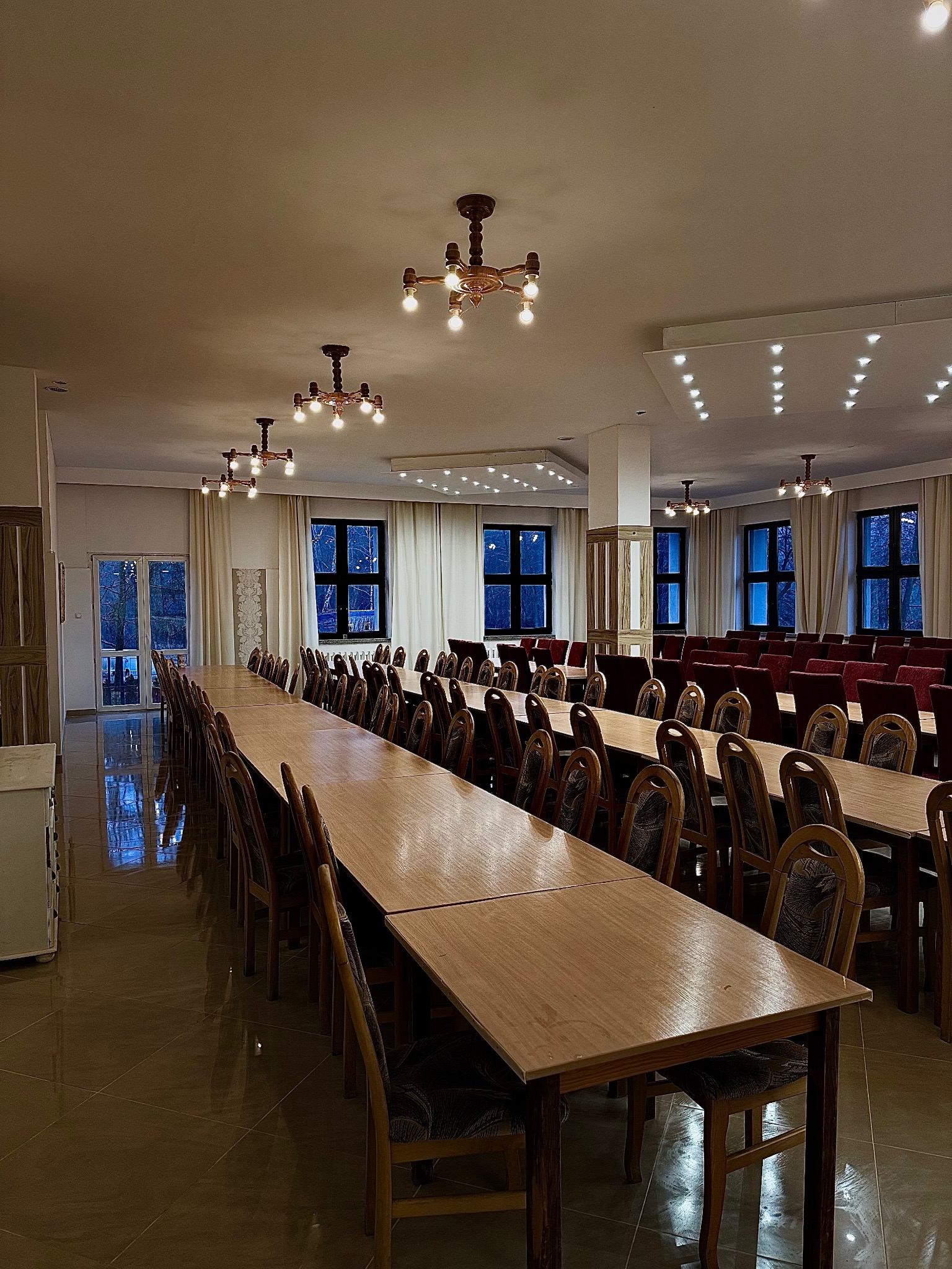 Sala bankietowa z długimi stołami i krzesłami, przygotowana do przyjęcia gości, oświetlona ozdobnymi żyrandolami, widok na okna z zasłonami.