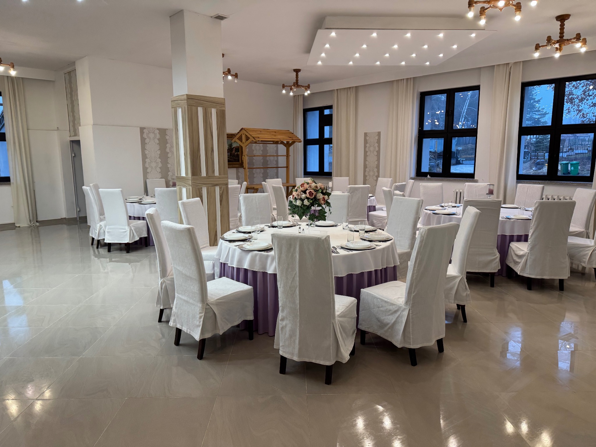 Sala bankietowa z okrągłymi stołami nakrytymi białymi obrusami z fioletowymi falbanami, krzesła w białych pokrowcach, drewniana dekoracja w tle, jasna podłoga z połyskiem.