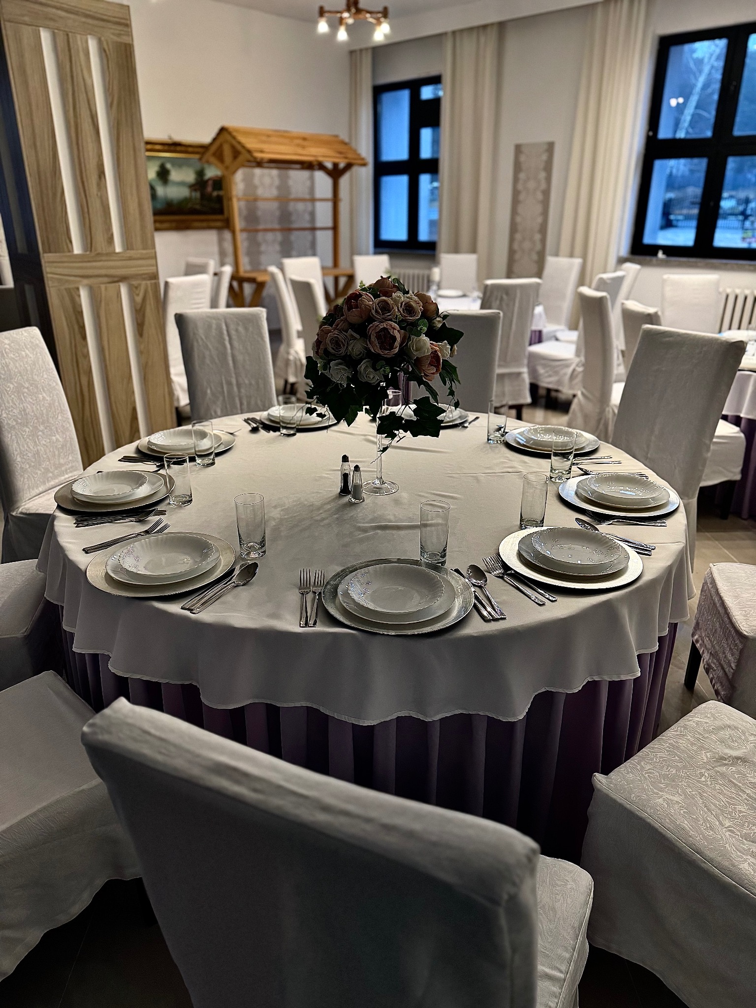 Elegancko nakryty okrągły stół z białym obrusem i fioletowymi akcentami, otoczony krzesłami w białych pokrowcach, w tle drewniana dekoracja i okna z ciemnymi ramami.