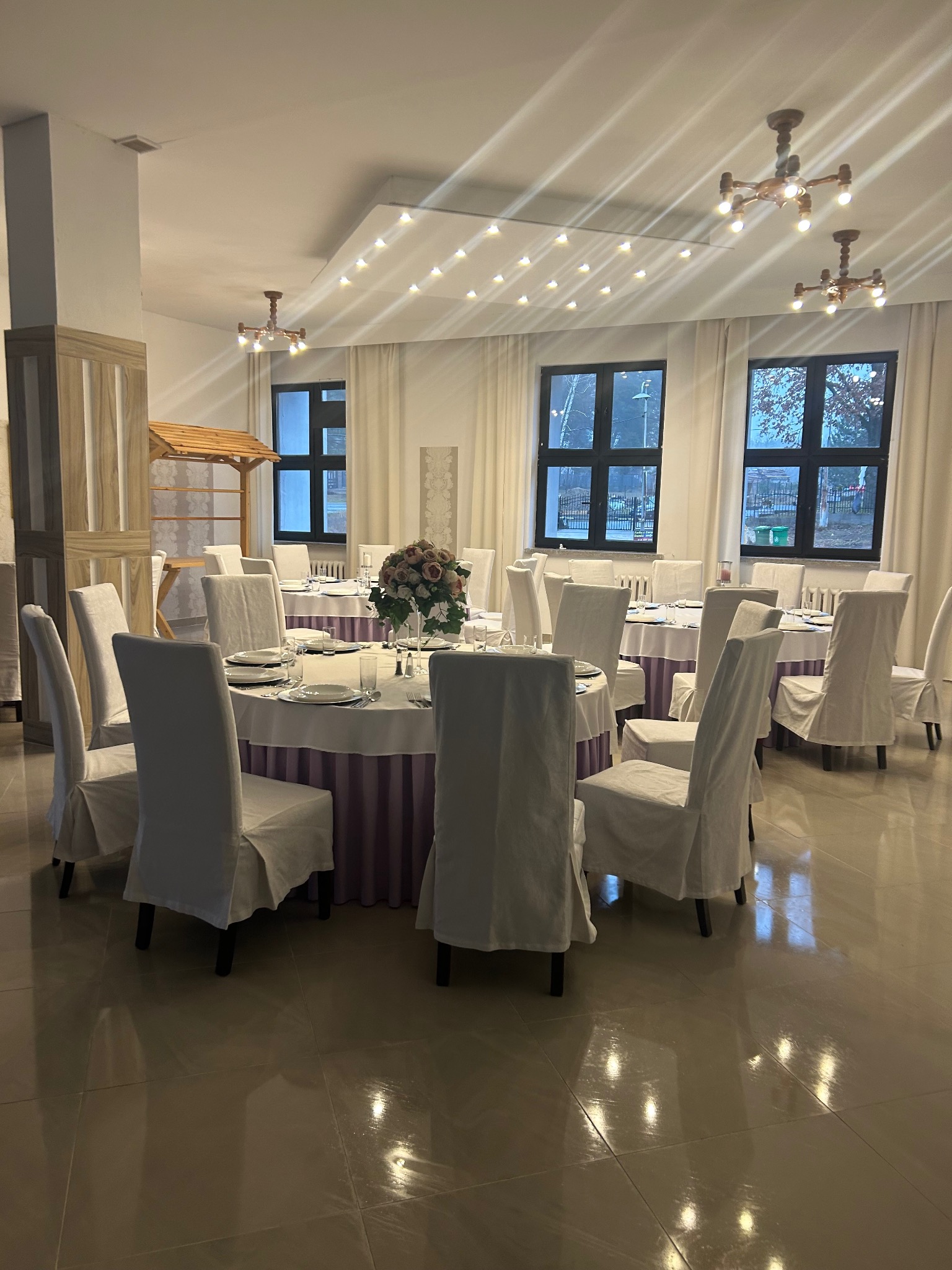 Elegancko nakryte okrągłe stoły z białymi obrusami i pokrowcami na krzesłach w jasnej sali bankietowej, dekoracja kwiatowa na stole, widok na okna i oświetlenie sufitowe.