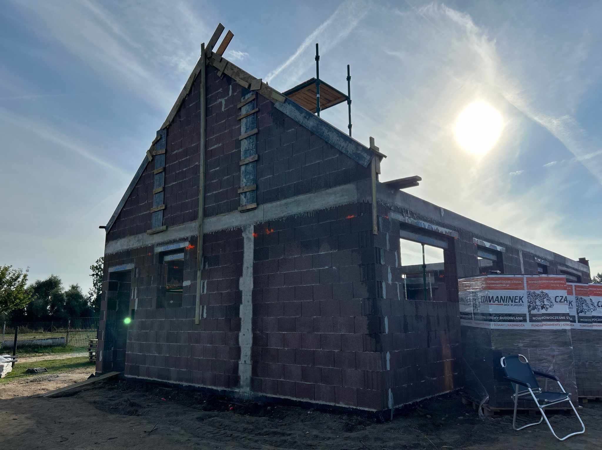 Surowy stan domu jednorodzinnego w trakcie budowy, ściany z bloczków, widoczne rusztowanie i materiały budowlane, jasne niebo w tle.
