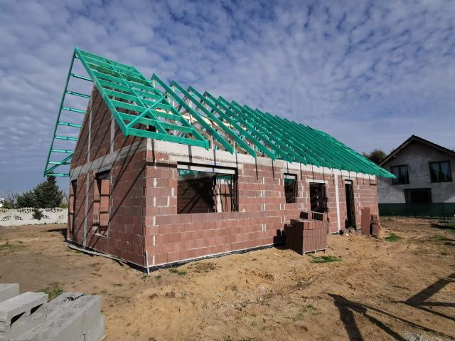 Dom w trakcie budowy: ściany z czerwonego bloczka, zielona konstrukcja dachu, widok na sąsiedni budynek w tle.