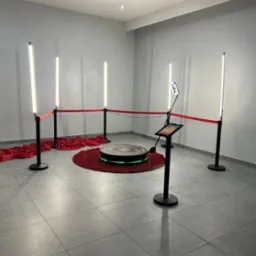 Oświetlona platforma obrotowa z ekranem dotykowym i obiektywem kamery otoczona czerwonym sznurem i czterema lampami LED, ułożona na czerwonym dywanie z dekoracyjną tkaniną, tworząca przestrzeń...