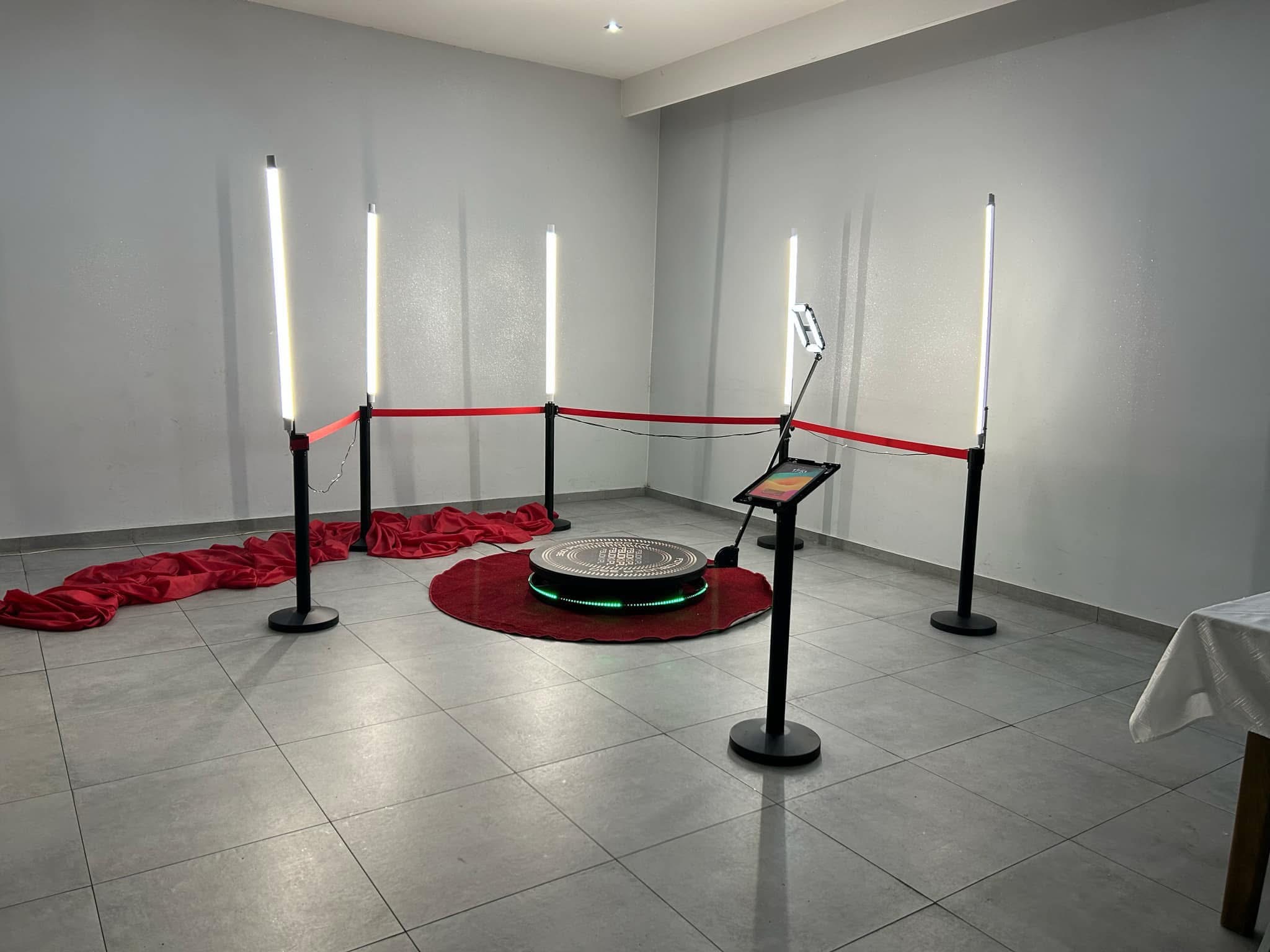 Oświetlona platforma obrotowa z ekranem dotykowym i obiektywem kamery otoczona czerwonym sznurem i czterema lampami LED, ułożona na czerwonym dywanie z dekoracyjną tkaniną, tworząca przestrzeń...