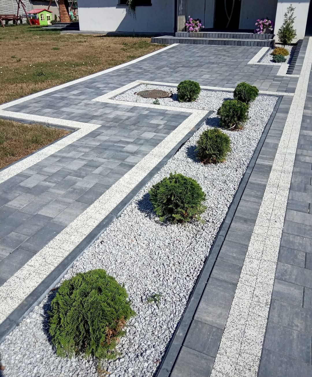 Szara kostka brukowa z białymi obrzeżami tworząca ścieżkę i podjazd. Dekoracyjne rabaty z białym grysem i kulistymi krzewami. Widok na dom z zielonym trawnikiem.