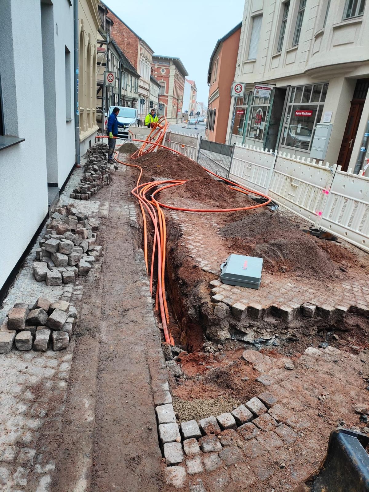 Ulica w trakcie prac: wykop z pomarańczowymi rurami, kostka brukowa w stosach i częściowo ułożona. Widoczni robotnicy i zabezpieczenia. Prace ziemne w przestrzeni miejskiej.