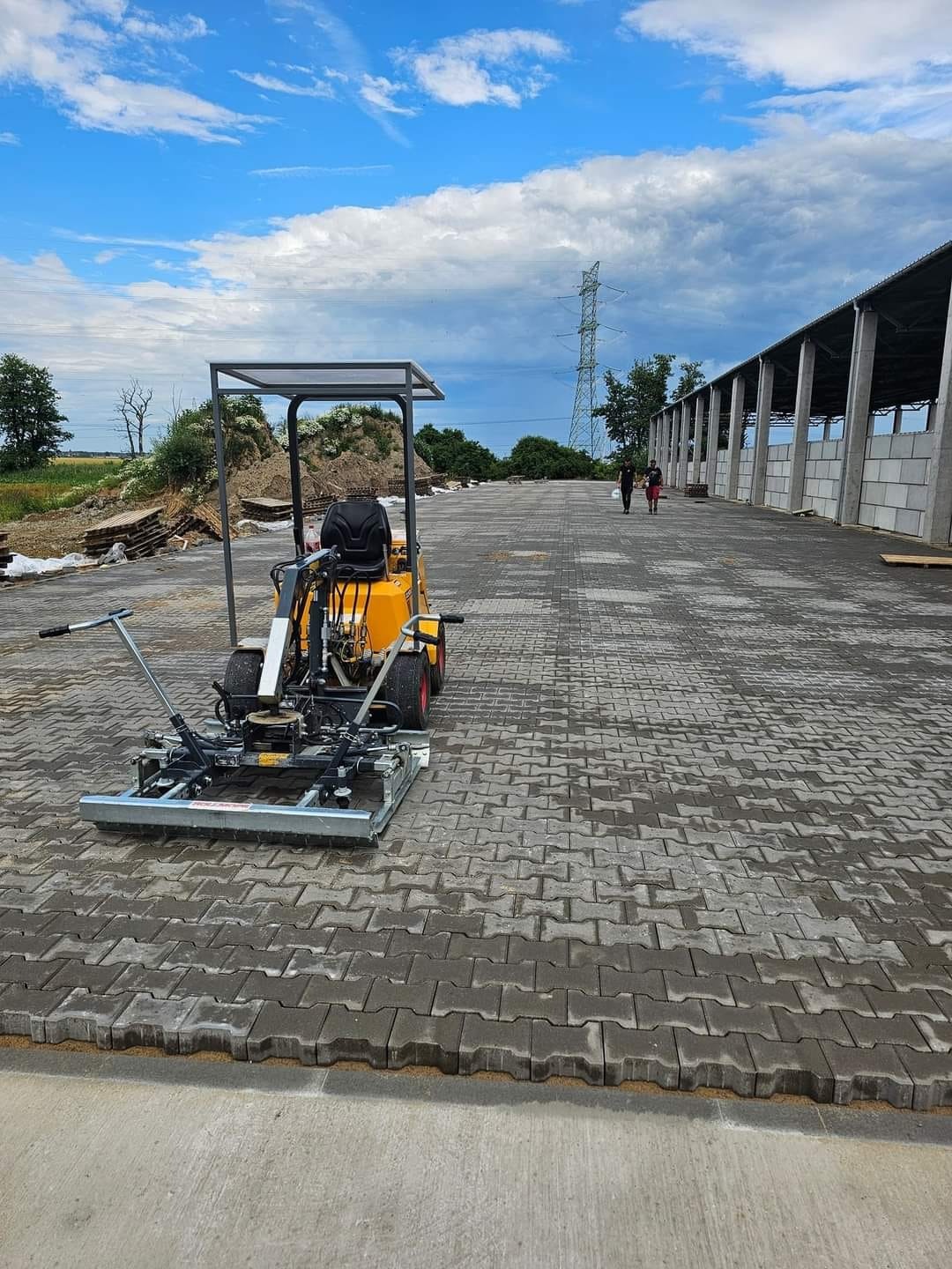Maszyna do układania kostki brukowej na dużej powierzchni, widoczne ułożone fragmenty, w tle budynek gospodarczy i słup wysokiego napięcia.