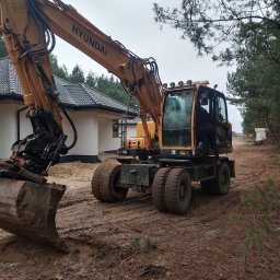 RAFTRANS Rafał Nejman - Żółta koparka kołowa Hyundai na błotnistym terenie budowy, operator w kabinie, w tle biały dom z ciemną dachówką i las.