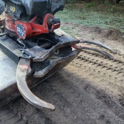 RAFTRANS Rafał Nejman - Ciężka, używana łyżka koparki z hydraulicznymi chwytakami, widoczne ślady użytkowania i ziemia, na piaszczystym podłożu z odciśniętym bieżnikiem opon.