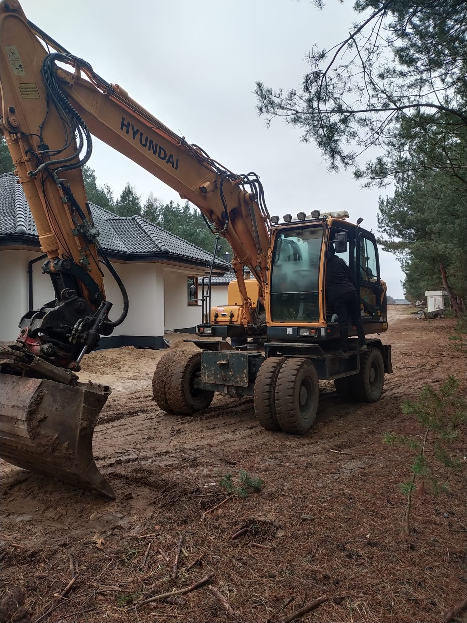 Żółta koparka kołowa Hyundai na błotnistym terenie budowy, operator w kabinie, w tle biały dom z ciemną dachówką i las.