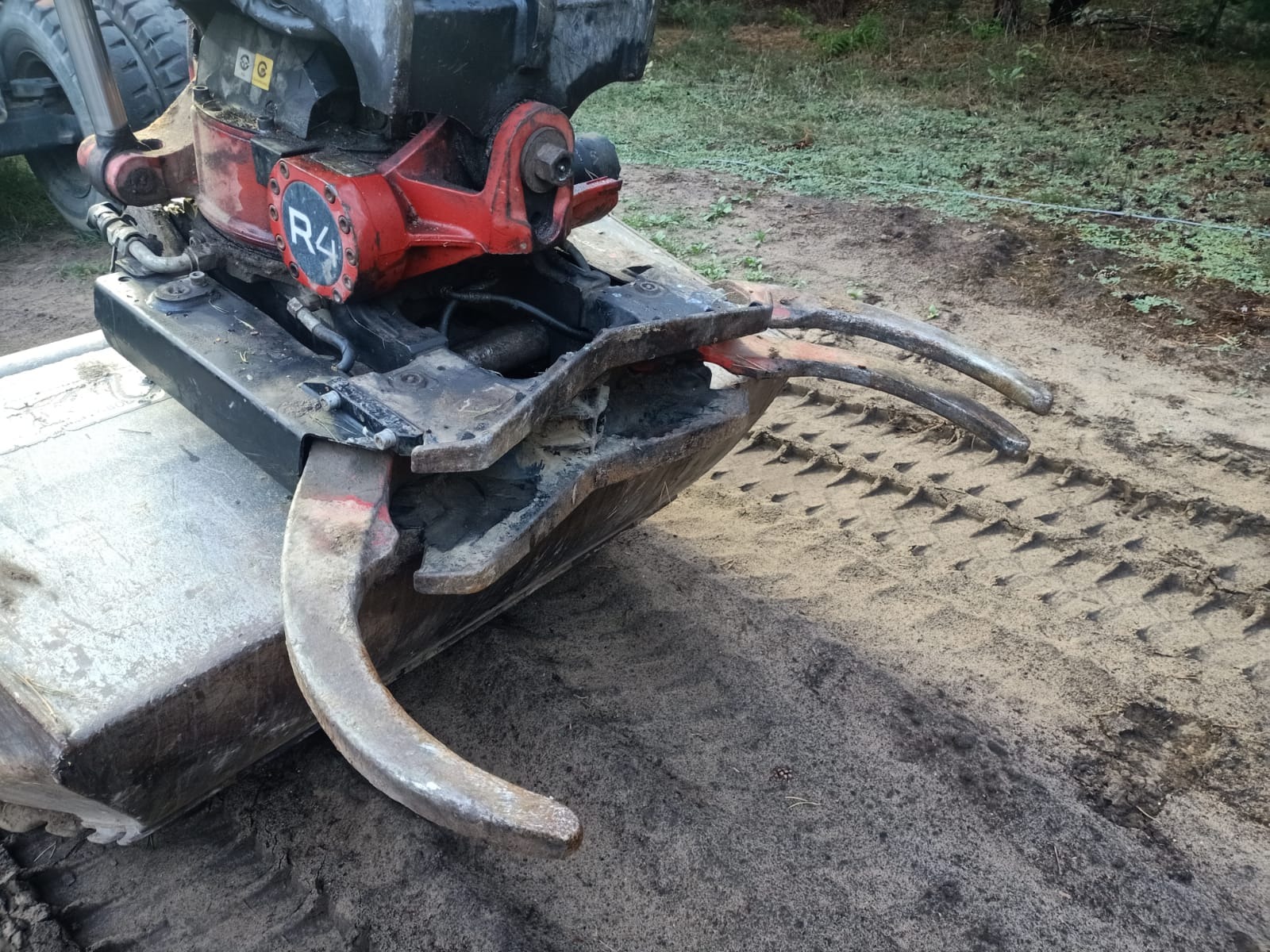 Ciężka, używana łyżka koparki z hydraulicznymi chwytakami, widoczne ślady użytkowania i ziemia, na piaszczystym podłożu z odciśniętym bieżnikiem opon.
