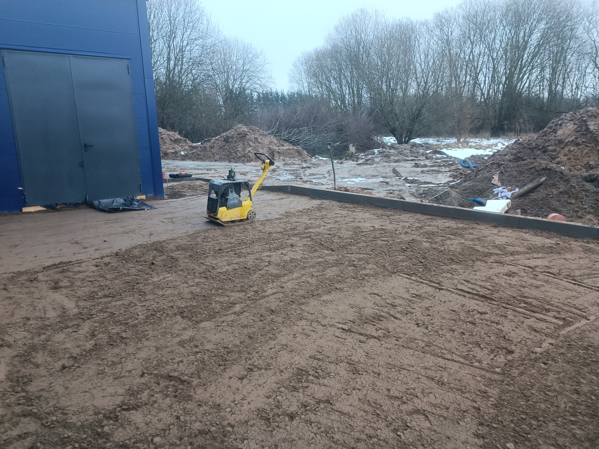 Żółta zagęszczarka spalinowa na ubitym gruncie, widoczne ślady pracy maszyny, w tle budynek z niebieską elewacją i hałdy ziemi z resztkami śniegu.