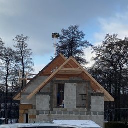 IKBUD - Wznoszenie domu z bloczków betonowych, widoczne drewniane elementy konstrukcji dachu, na szczycie maszt z wieńcem, w tle drzewa i pochmurne niebo, częściowo widoczny samochód.