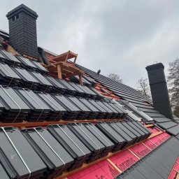 IKBUD - Układanie czarnej dachówki ceramicznej na dachu, widoczny komin z cegły i drewniany podest roboczy, zabezpieczenie membraną dachową.