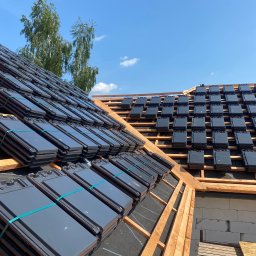 IKBUD - Układanie ciemnoszarej dachówki ceramicznej na konstrukcji drewnianej dachu, widoczne pakiety dachówek zabezpieczone zieloną taśmą, na tle błękitnego nieba i drzew