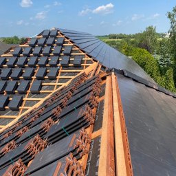 IKBUD - Częściowo pokryty dach z drewnianą konstrukcją, widoczne czarne dachówki ceramiczne, niektóre jeszcze w paczkach, oraz fragmenty już ułożone na dachu, w tle zieleń i błękitne niebo.