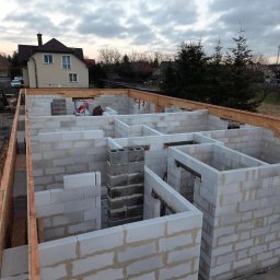 IKBUD - Wznoszenie ścian parteru z betonu komórkowego Ytong, widoczny komin z szarych bloczków oraz drewniane szalunki stropu, w tle dom jednorodzinny i pochmurne niebo.
