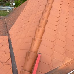 IKBUD - Układanie ceramicznej dachówki karpiówki w kolorze ceglastym, widoczny fragment kalenicy i poziomica, praca w toku.