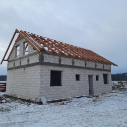 IKBUD - Niedokończony dom murowany z białych bloczków w zimowej scenerii, z drewnianą konstrukcją dachu bez pokrycia, widoczne stosy drewna i śnieg na ziemi.