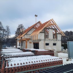 IKBUD - Surowy stan domu murowanego z drewnianą konstrukcją dachu w zimowej scenerii, palety czerwonej dachówki pokryte śniegiem na pierwszym planie.