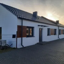 Szeregowy dom murowany z białymi ścianami, ciemną dachówką i elementami dekoracyjnymi z drewna, widok z zewnątrz na tle zachodzącego słońca.