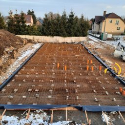 IKBUD - Zbrojenie płyty fundamentowej z rurami instalacyjnymi, widok z góry na plac budowy w zimowej scenerii.