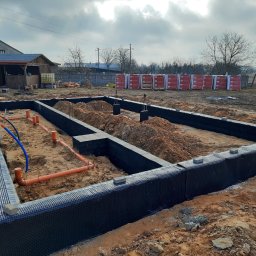 IKBUD - Przygotowany wykop pod fundamenty z izolacją pionową, widoczne rury kanalizacyjne, w tle palety z materiałami budowlanymi i budynek gospodarczy.