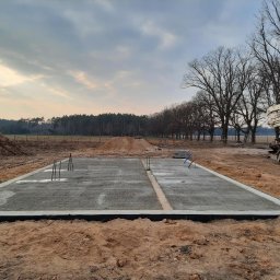 IKBUD - Wylany fundament z wystającymi prętami zbrojeniowymi, widoczny betonowóz na tle drzew i zachmurzonego nieba.