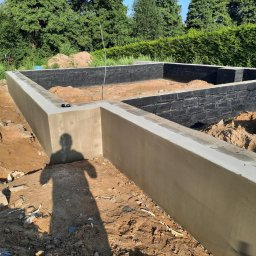 IKBUD - Zabezpieczone fundamenty domu jednorodzinnego, ściany pomalowane na czarno, teren wokół zasypany ziemią, cień osoby fotografującej widoczny na pierwszym planie.