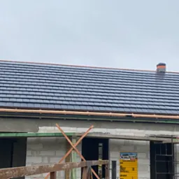 Budowa domu: fragment dachu pokrytego ciemną dachówką ceramiczną, komin owinięty folią ochronną, surowy stan elewacji z widocznymi bloczkami i drewnianym rusztowaniem.