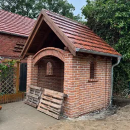 Murowana altana z czerwoną dachówką i drewnianą konstrukcją dachu. Widoczne palety obok konstrukcji. Tło stanowi zieleń drzew i krzewów.