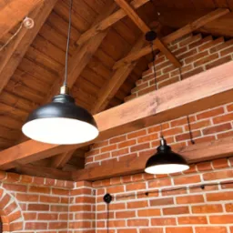 Ceglana ściana z drewnianą więźbą dachową, oświetlona dwoma czarnymi lampami wiszącymi. Widoczne detale instalacji elektrycznej i dekoracyjne oświetlenie.