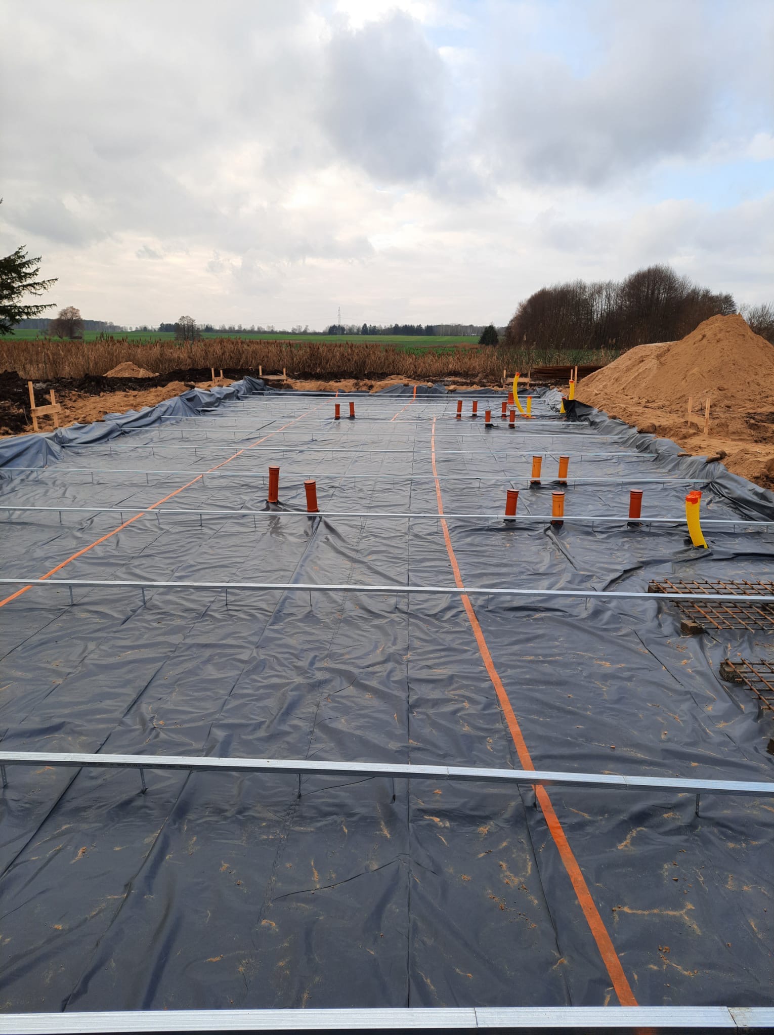Przygotowanie terenu pod fundamenty: rozłożona czarna folia hydroizolacyjna z zaznaczonymi pomarańczowymi liniami, wystające rury kanalizacyjne i stalowe szalunki, w tle hałda piasku i zarośla...