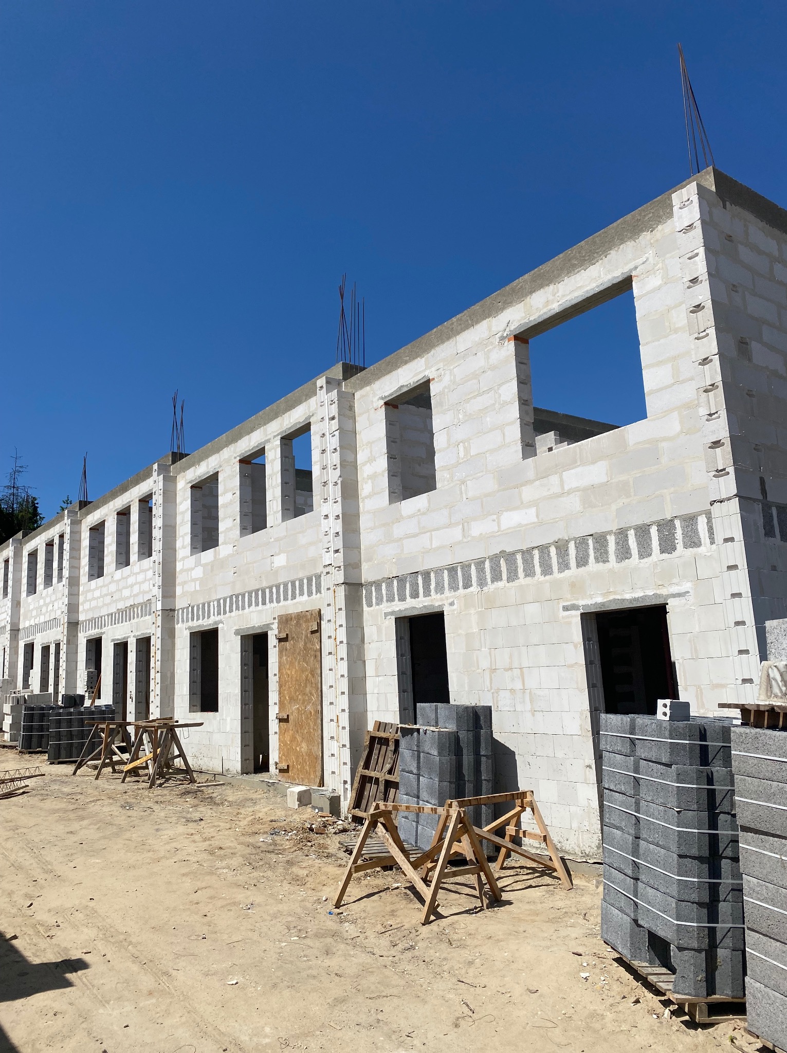 Wznoszenie ścian parterowych budynków z betonu komórkowego, widoczne otwory okienne i drzwiowe, szalunki oraz zapas bloczków na paletach, teren budowy w słoneczny dzień.