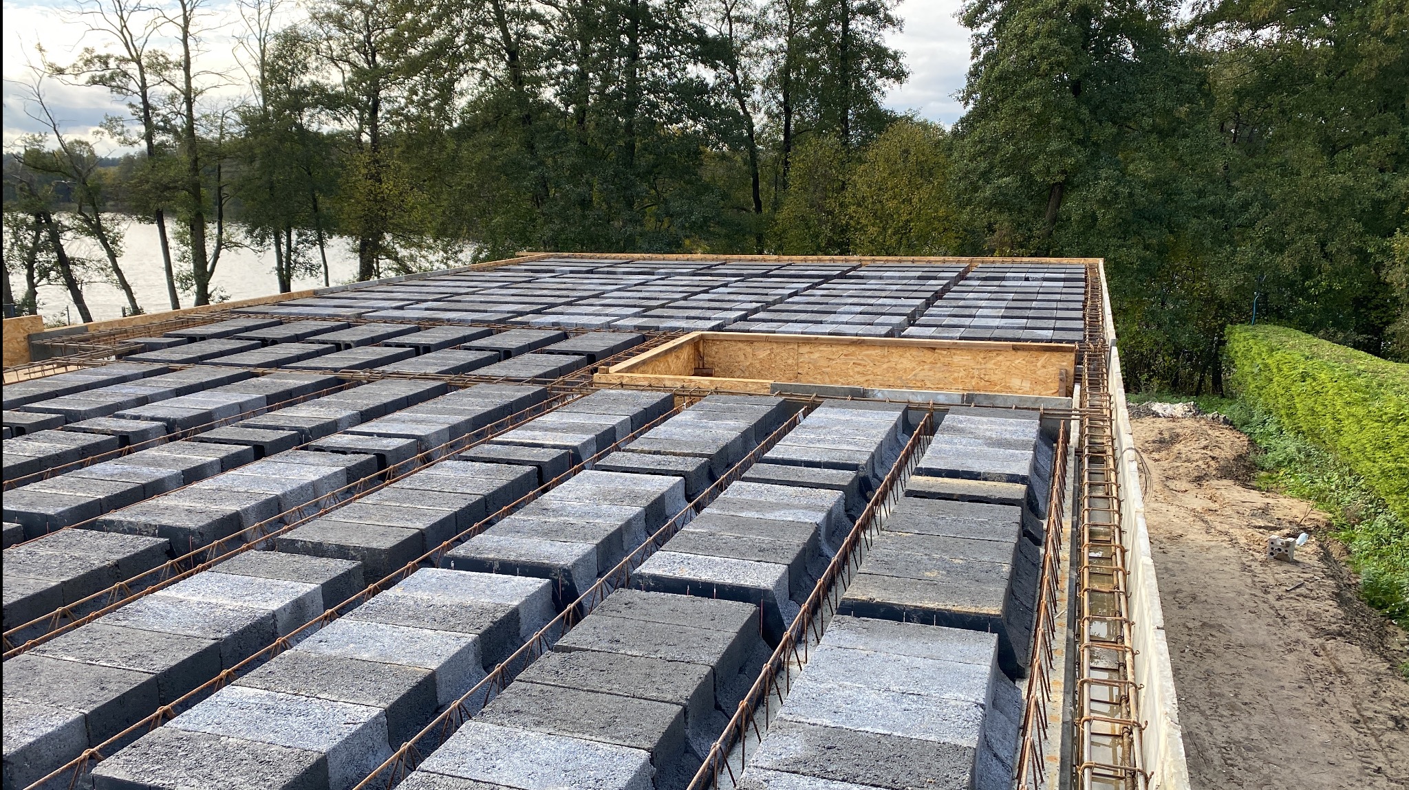 Wieniec stropowy w trakcie budowy, zbrojenie i betonowe pustaki szalunkowe na tle zieleni i jeziora.