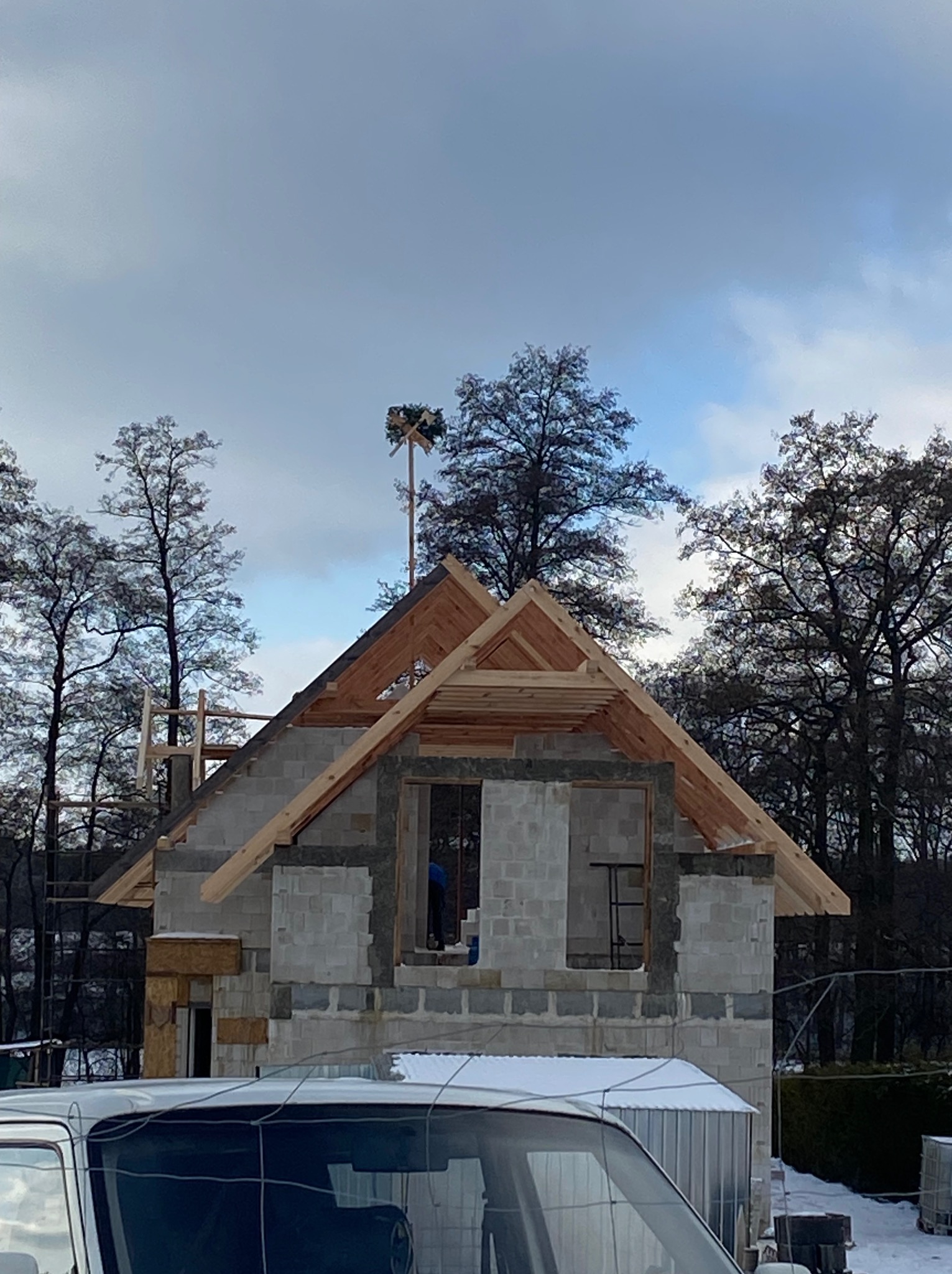 Wznoszenie domu z bloczków betonowych, widoczne drewniane elementy konstrukcji dachu, na szczycie maszt z wieńcem, w tle drzewa i pochmurne niebo, częściowo widoczny samochód.