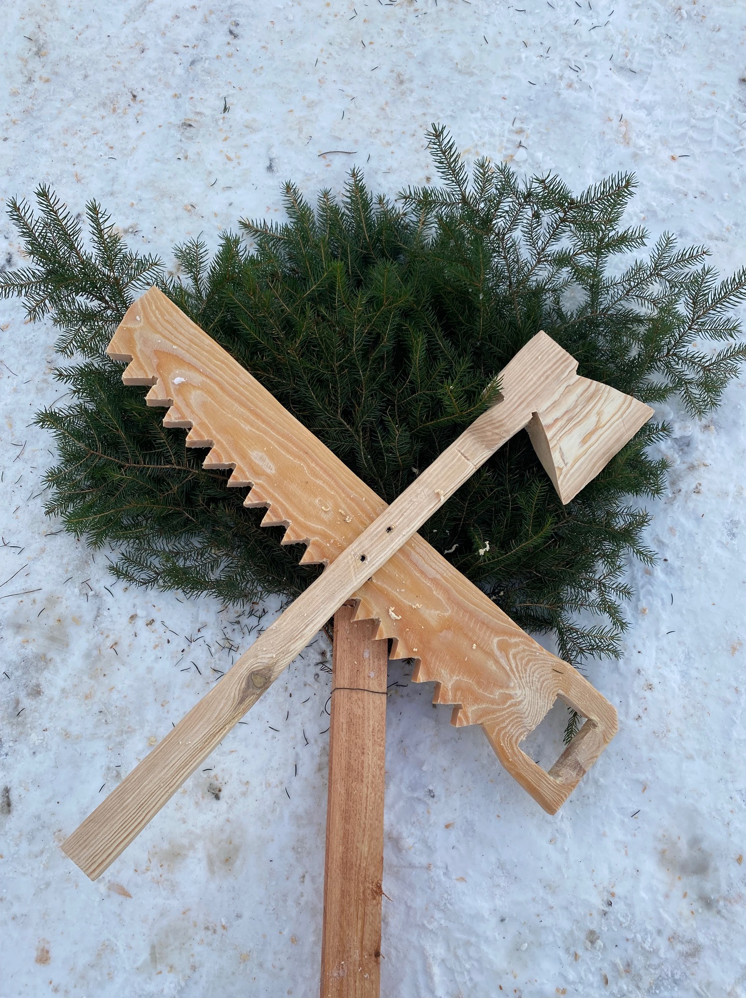 Dekoracja zimowa: drewniana piła i topór skrzyżowane na tle gałęzi świerkowych, leżące na śniegu.