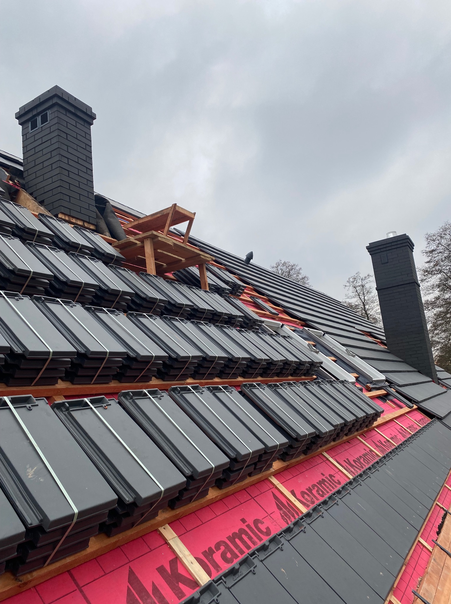 Układanie czarnej dachówki ceramicznej na dachu, widoczny komin z cegły i drewniany podest roboczy, zabezpieczenie membraną dachową.