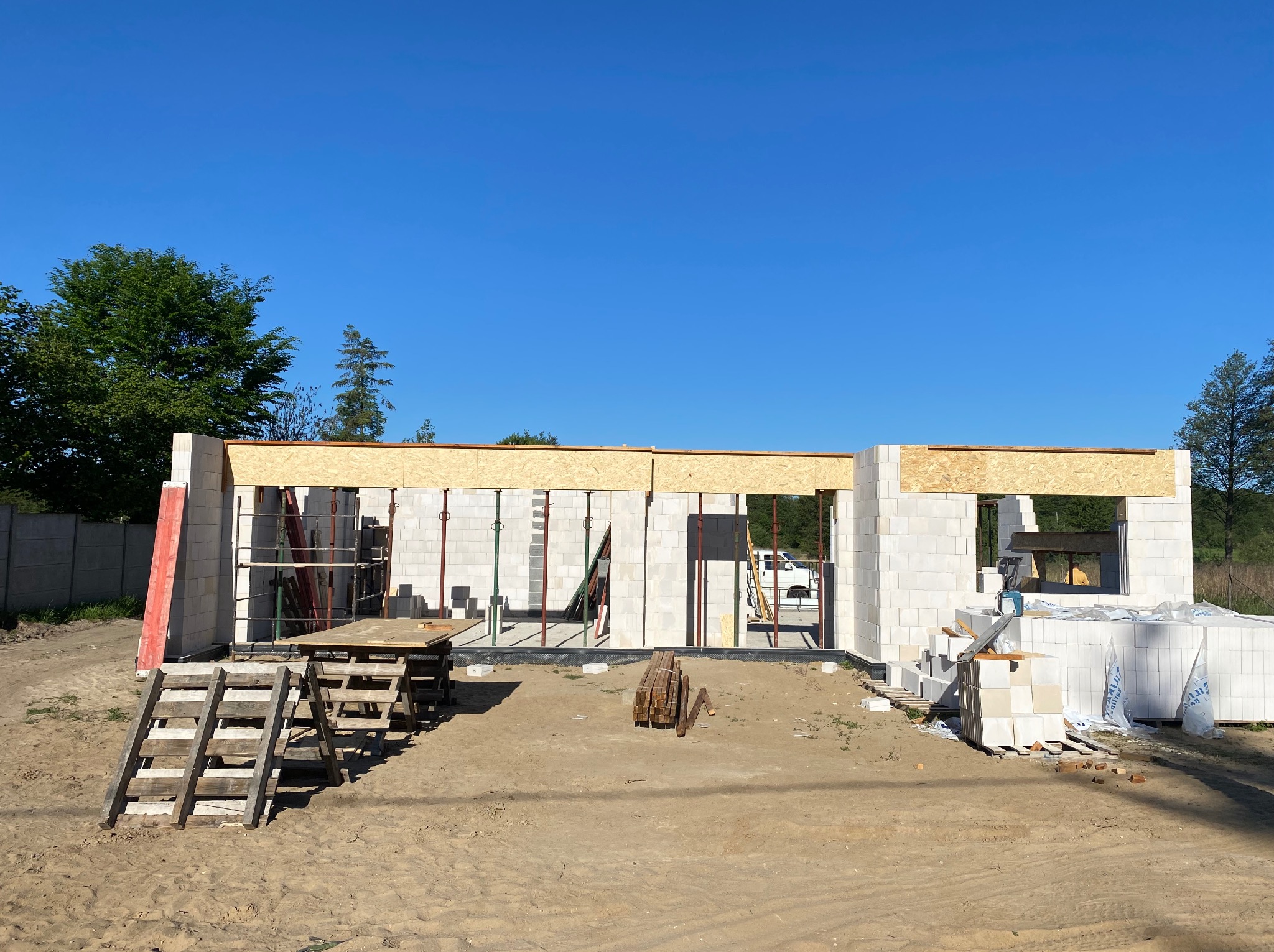 Wznoszenie ścian domu z bloczków betonu komórkowego, widoczne drewniane szalunki nad otworami okiennymi i drzwiowymi, w tle błękitne niebo i zieleń drzew.