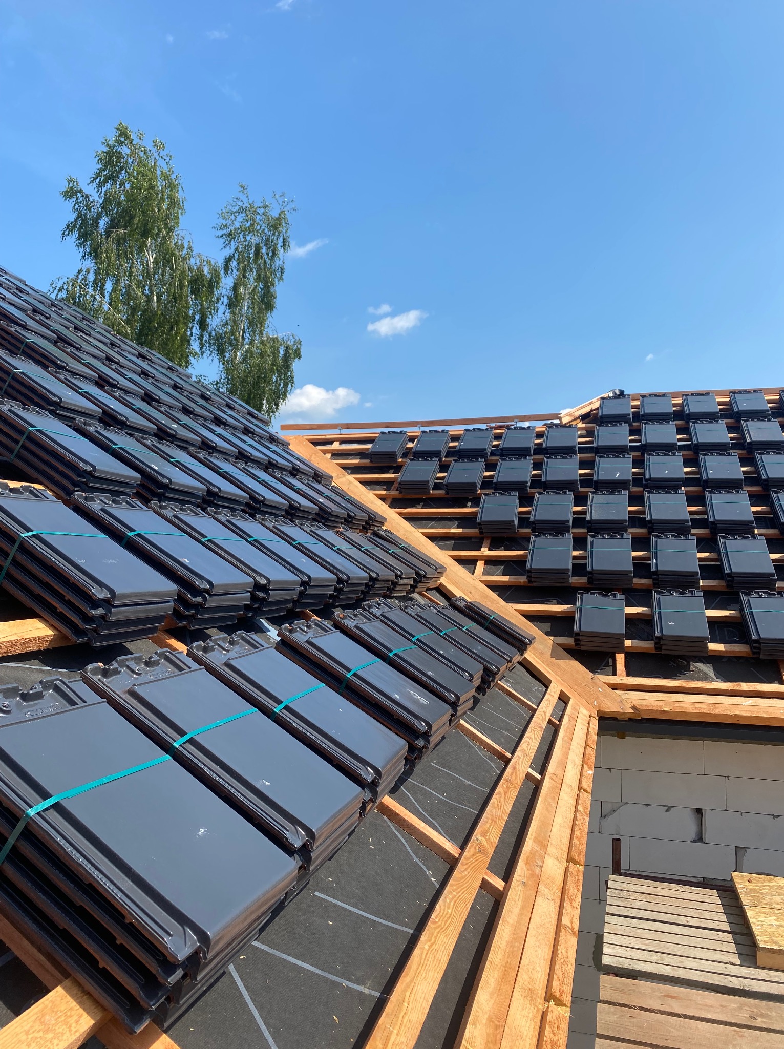 Układanie ciemnoszarej dachówki ceramicznej na konstrukcji drewnianej dachu, widoczne pakiety dachówek zabezpieczone zieloną taśmą, na tle błękitnego nieba i drzew